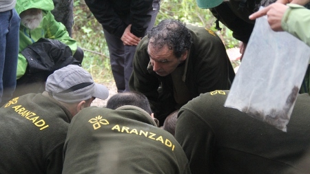  Aranzadis tvärvetenskapliga team står på knä och gräver sig försiktigt fram genom leran. Fynden dokumenteras noggrant och läggs i bevispåsar.
